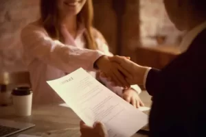Jak dostać pracę jako rekruter? Kobieta i mężczyzna podają sobie rękę nad biurkiem. Mężczyzna trzyma umowę w ręku.